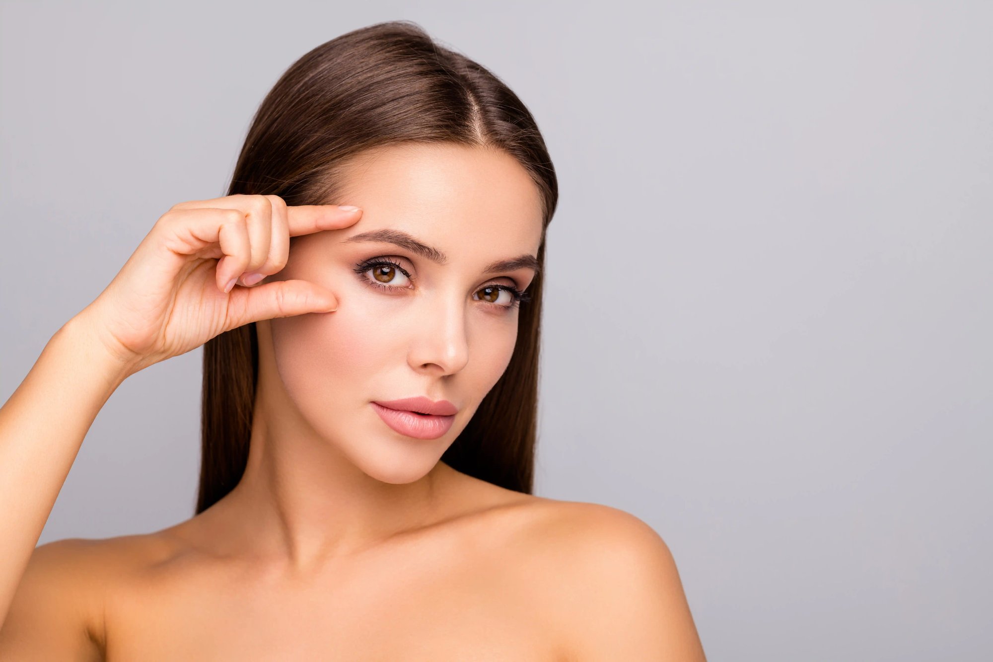 Illustrative stock photo of a woman lifting her eyebrow with a fingertip against a grey studio background; not a Leva patient