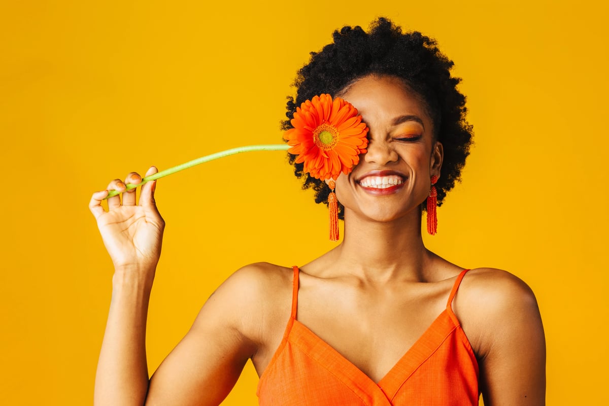 Illustrative stock photo of a smiling woman holding an orange gerbera daisy against a yellow studio background; not a Leva patient